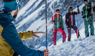Eine Gruppe von Menschen in farbenfroher Schneeausrüstung steht auf einem schneebedeckten Hang. Eine Person im Vordergrund hält einen Skistock und gestikuliert, wahrscheinlich um den anderen Anweisungen zu geben. Im Hintergrund sind schneebedeckte Berge zu sehen.
