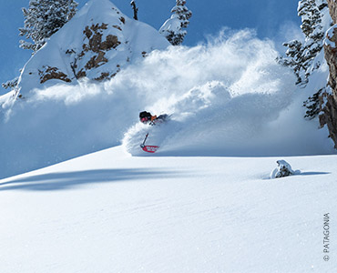 Ein Snowboarder in einer roten Jacke fährt durch den frischen Pulverschnee auf einem schneebedeckten Berghang und hinterlässt eine Spur. Im Hintergrund sind Kiefern und Felsvorsprünge unter einem klaren blauen Himmel zu sehen.