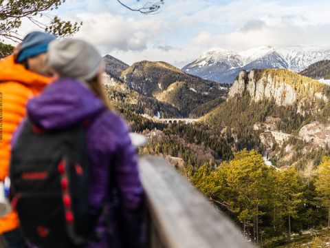 Zwei Menschen in warmen Jacken stehen auf einer Holzplattform und blicken auf eine malerische Berglandschaft. Schneebedeckte Gipfel und dichte Wälder erstrecken sich über die Aussicht, in der Ferne ist ein Steinviadukt zu sehen. Die Szene spielt unter einem hellen, teilweise bewölkten Himmel.