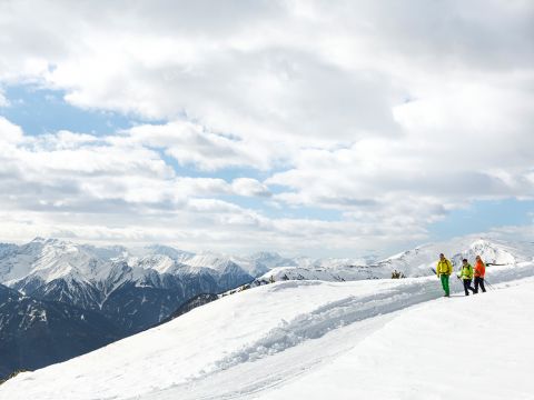 Drei Menschen gehen unter einem bewölkten Himmel auf einem schneebedeckten Bergpfad. Um sie herum sind ausgedehnte Bergketten mit schneebedeckten Gipfeln. Die Szene fängt die ruhige, kalte Atmosphäre einer Hochgebirgslandschaft ein.