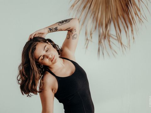 Eine Frau in einem schwarzen Tanktop posiert mit geneigtem Kopf und berührt mit der Hand ihr Haar. Sie hat Tattoos auf dem Arm. In der oberen rechten Ecke ist vor einem hellen Hintergrund ein getrocknetes Palmblatt zu sehen.