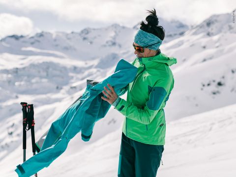 Eine Person in leuchtend grüner und blauer Winterkleidung steht auf einem schneebedeckten Berghang und hält eine Jacke in der Hand. Sie trägt ein Stirnband und eine Sonnenbrille, im Hintergrund sind Skistöcke zu sehen. Unter einem bewölkten Himmel sind schneebedeckte Gipfel zu sehen.