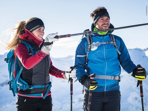 Zwei Menschen in Winterkleidung stehen lächelnd auf einem schneebedeckten Berg. Sie halten Skistöcke in der Hand, im Hintergrund sind schneebedeckte Gipfel zu sehen. Der Himmel ist klar und hell, was auf einen sonnigen Tag in den Bergen schließen lässt.