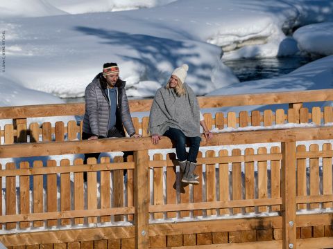 Ein Mann und eine Frau, beide in Winterkleidung, lehnen an einem Holzzaun einer schneebedeckten Brücke. Sie lächeln und schauen einander an, umgeben von einer verschneiten Landschaft.