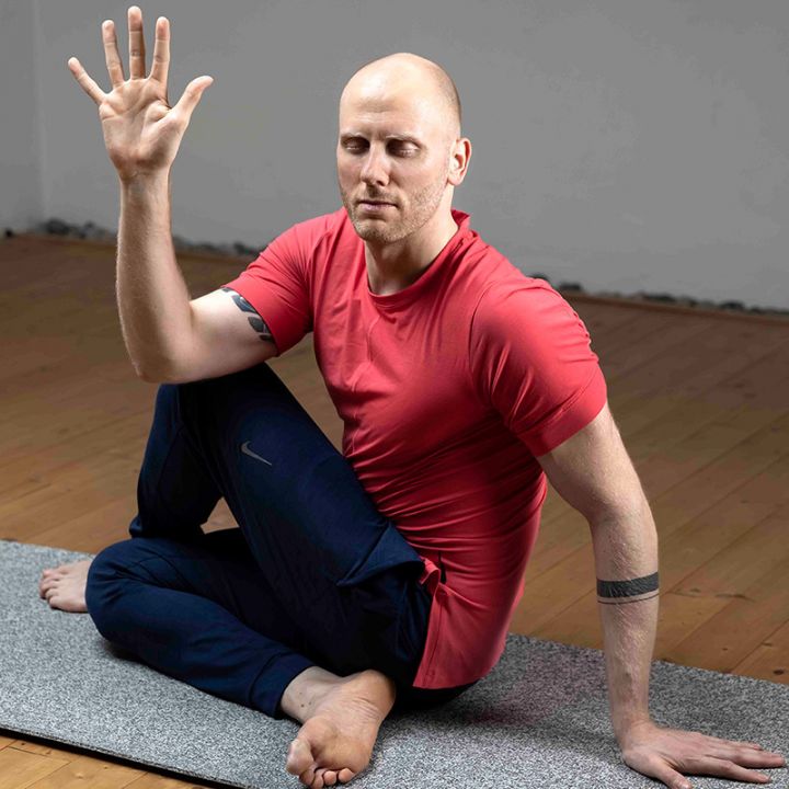 Eine Person in einem roten Hemd und schwarzen Hosen sitzt auf einer Yogamatte und führt eine Drehpose aus. Sie sitzt im Schneidersitz, ein Arm ist erhoben und der andere ruht auf dem Knie. Der Hintergrund ist ein schlichter Holzboden und eine graue Wand.