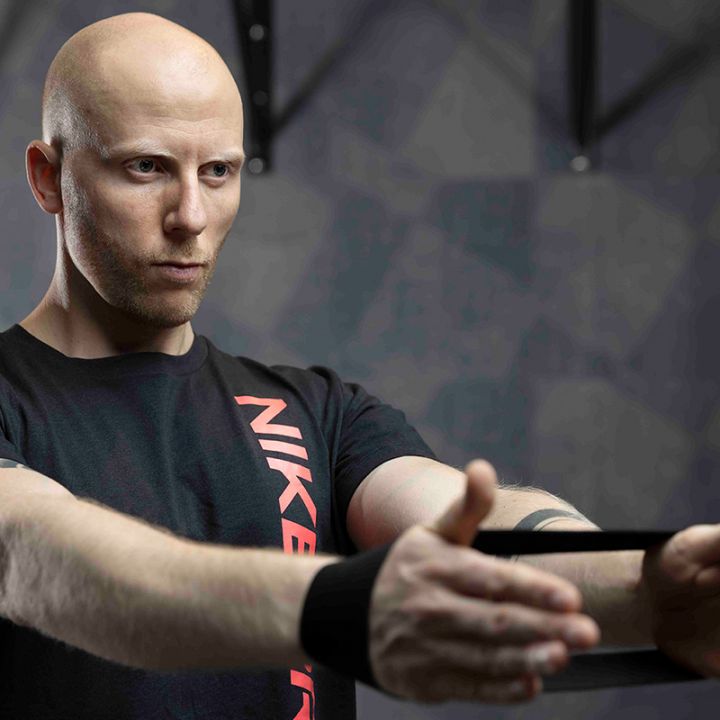 Ein kahlköpfiger Mann in einem schwarzen T-Shirt mit roter Schrift spannt ein Widerstandsband zwischen seinen Händen und steht mit konzentriertem Gesichtsausdruck in einem Fitnessstudio. Der Hintergrund ist mit geometrischen Mustern verschwommen.
