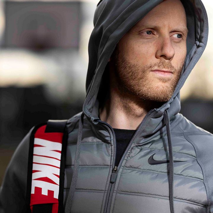 Ein Mann in einer grauen Kapuzenjacke steht mit nachdenklichem Gesichtsausdruck im Freien. Er trägt eine Sporttasche mit einem sichtbaren „NIKE“-Logo. Der Hintergrund ist verschwommen und suggeriert eine Sportkulisse. Ein Basketballkorb ist schwach zu erkennen.