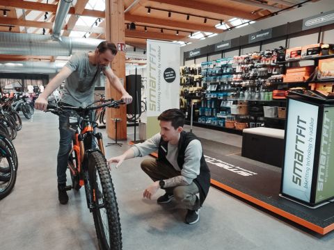 Zwei Personen in einem Fahrradladen: Eine hockt und inspiziert das Hinterrad eines orange-schwarzen Fahrrads, die andere steht neben dem Rad. Im Hintergrund sind Regale mit Fahrradzubehör zu sehen.