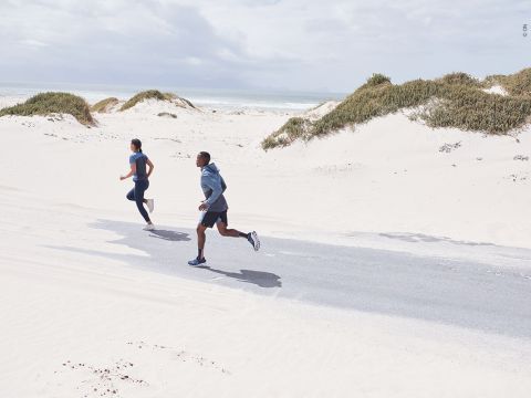Zwei Personen laufen auf einer asphaltierten Straße durch eine sandige Strandlandschaft. Sie tragen Sportkleidung und der Himmel ist teilweise bewölkt. Um sie herum sind Sanddünen und spärliche Vegetation zu sehen.
