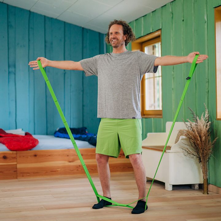 Eine Person in einem grauen T-Shirt und grünen Shorts steht lächelnd in einem Raum und spannt mit ausgestreckten Armen ein grünes Widerstandsband. Der Raum hat Holzwände und -böden, Fenster und ein Bett mit roter und blauer Bettwäsche.