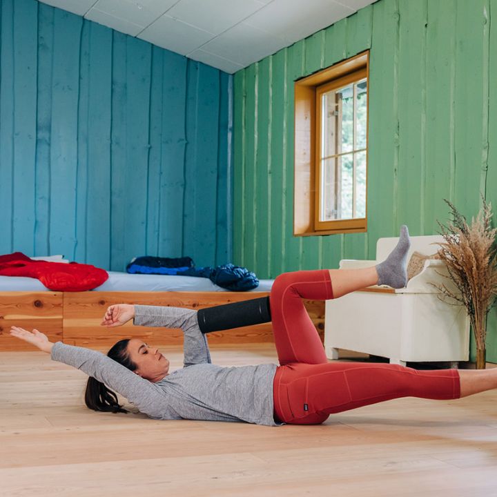 Eine Person in roten Leggings und einem grauen Langarmshirt führt auf einem Holzboden in einem Raum mit grünen Wänden und zwei Fenstern eine Rumpfübung durch. Ein Arm und das gegenüberliegende Bein sind angehoben, während der andere Arm und das andere Bein auf dem Boden bleiben.