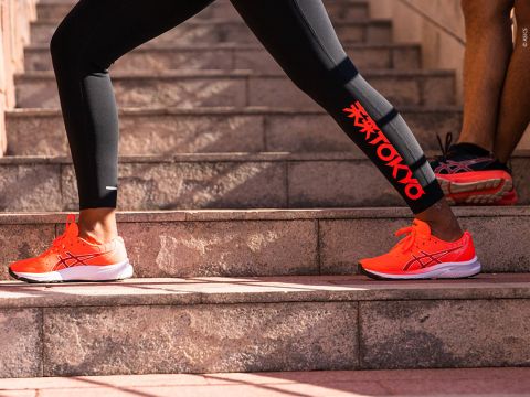 Eine Person trägt schwarze Leggings mit rotem „Tokio“-Aufdruck und leuchtend orangefarbene Laufschuhe und macht Dehnübungen auf Steinstufen. Eine andere Person in Shorts und passenden Schuhen steht in der Nähe und präsentiert Sportschuhe und Sportausrüstung.