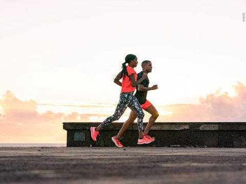 Zwei Menschen joggen nebeneinander bei Sonnenaufgang oder Sonnenuntergang; einer trägt ein helles Shirt und gemusterte Leggings, der andere ein einfarbiges Outfit. Beide haben Laufschuhe an. Der Himmel ist mit weichen Wolken und warmen Farbtönen gefüllt.