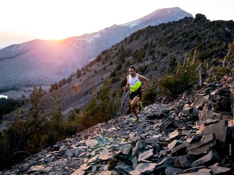 Eine Person in Sportkleidung läuft bei Sonnenaufgang einen felsigen Bergpfad hinauf. Die Sonne geht über dem Horizont auf und taucht die Landschaft in einen warmen Glanz. Im Hintergrund sind hohe Bäume und Berggipfel zu sehen.