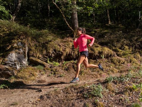 Eine Person in einem roten Langarmshirt und schwarzen Shorts läuft energisch einen Waldweg entlang. Der Boden ist uneben und von dichtem Grün und Bäumen umgeben, durch deren Blätter Sonnenlicht fällt.