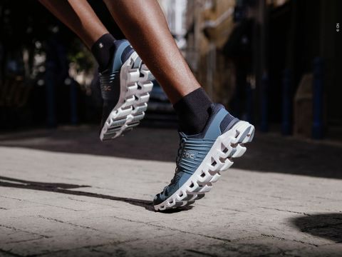 Nahaufnahme einer Person, die auf einer Straße läuft. Sie trägt blau-weiße Sportschuhe mit einzigartigen, wolkenartigen Sohlen. Der Fokus liegt auf dem Schuhwerk, das den Läufer mitten im Schritt einfängt und Bewegung und Beweglichkeit hervorhebt.