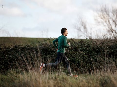 Eine Person läuft im Freien auf einer Wiese bei bewölktem Himmel. Sie trägt ein grünes Langarmshirt, schwarze Leggings und rote Laufschuhe. Im Hintergrund sind blattlose Bäume und eine Hecke zu sehen.