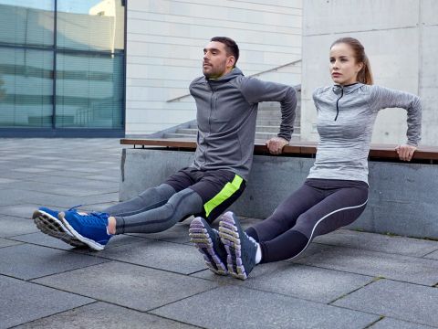 Zwei Personen in Sportkleidung machen Trizeps-Dips auf einer Bank im Freien. Sie sind konzentriert und halten ihre Beine gestreckt, die Füße stehen auf dem Boden. Im Hintergrund ist ein modernes Gebäude mit großen Glasfenstern zu sehen.