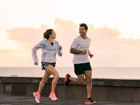 Ein Mann und eine Frau joggen nebeneinander auf einer gepflasterten Fläche in der Nähe des Meeres. Sie tragen Sportkleidung, die Frau trägt eine leichte Jacke und helle Turnschuhe. Sie lächeln sich an, im Hintergrund sind ein bewölkter Himmel und das Meer zu sehen.