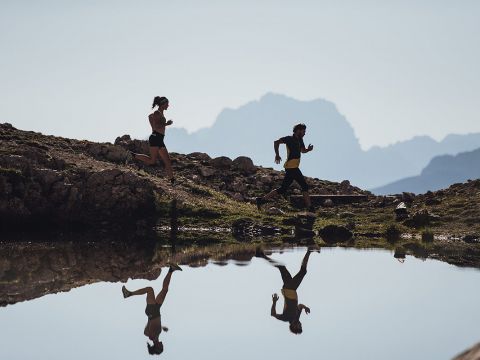 Zwei Menschen laufen auf einem steinigen Pfad neben einem ruhigen Gewässer entlang, in dem sich ihre Silhouetten spiegeln. Im Hintergrund ist eine dunstige Bergkette unter einem klaren Himmel zu sehen.