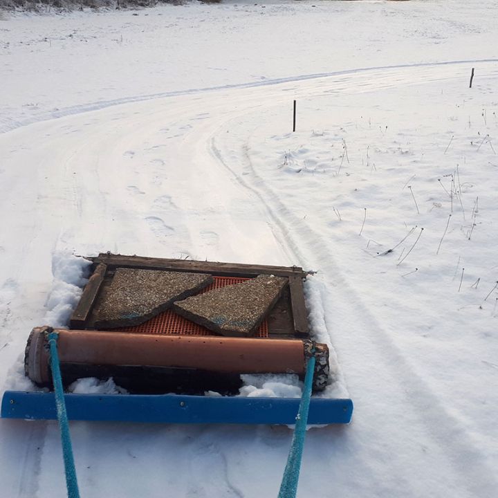 Auf einem verschneiten Weg ist ein Pistenraupengerät zu sehen, dessen Reifenspuren in die Ferne führen. Das Gerät ist rechteckig mit Teppichabschnitten und einem Gitter, das von blauen Seilen gezogen wird. Auf der rechten Seite ist spärliche Vegetation zu sehen.