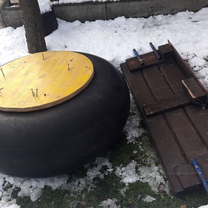 Ein großer schwarzer Reifen mit einem gelben Holzbrett darauf liegt auf schneebedecktem Gras neben einem zerlegten Holztisch mit blauen Klammern. In der Nähe ist ein Baumstamm zu sehen.