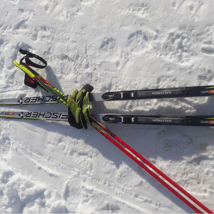 Zwei Paar Langlaufski und Stöcke liegen auf einem schneebedeckten Boden. Die Ski sind überwiegend schwarz mit grünen und roten Akzenten, und die Stöcke sind rot. Der Schnee zeigt Spuren und Fußabdrücke rund um die Ausrüstung.