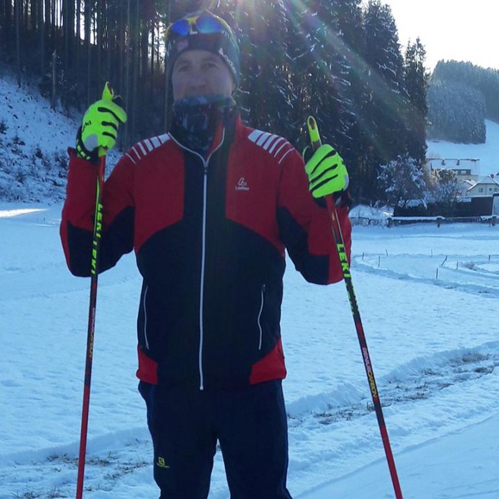 Eine Person in rot-schwarzem Winteroutfit steht mit Skistöcken im Freien auf dem Schnee und ist bereit zum Skifahren. Im Hintergrund sind Bäume und ein Haus zu sehen. Die Sonne scheint hell und wirft einen Schein auf die Schneeoberfläche.