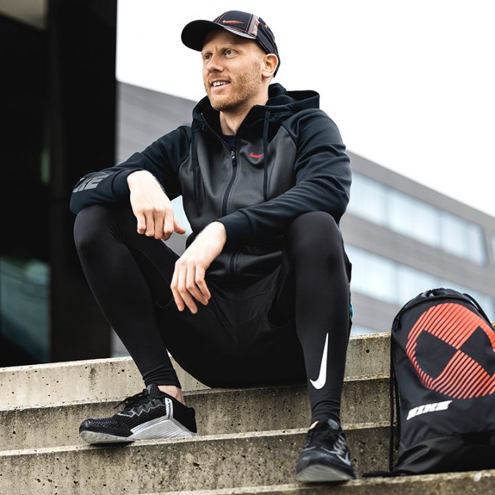 Eine Person in schwarzer Sportkleidung und einer Kappe sitzt auf Betonstufen im Freien. Sie lächelt und blickt zur Seite. Neben ihr steht eine Sporttasche mit einem geometrischen Muster. Im Hintergrund sind moderne Gebäudestrukturen zu sehen.