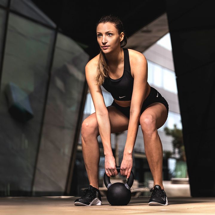 Eine Frau in Sportkleidung steht im Freien in einer städtischen Umgebung und hält mit beiden Händen eine Kettlebell. Sie sitzt in der Hocke und sieht konzentriert aus. Im Hintergrund sind moderne architektonische Elemente mit eckigen Linien zu sehen.