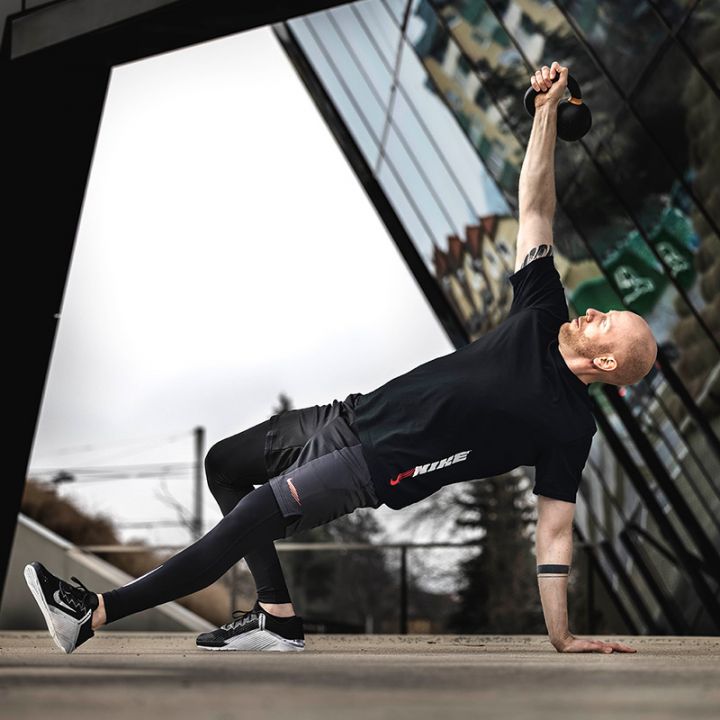 Eine Person in Sportkleidung führt vor einem modernen Gebäude mit reflektierendem Glas eine Kettlebell-Übung durch. Sie balanciert auf einer Hand und einem Fuß und streckt die Kettlebell nach oben.