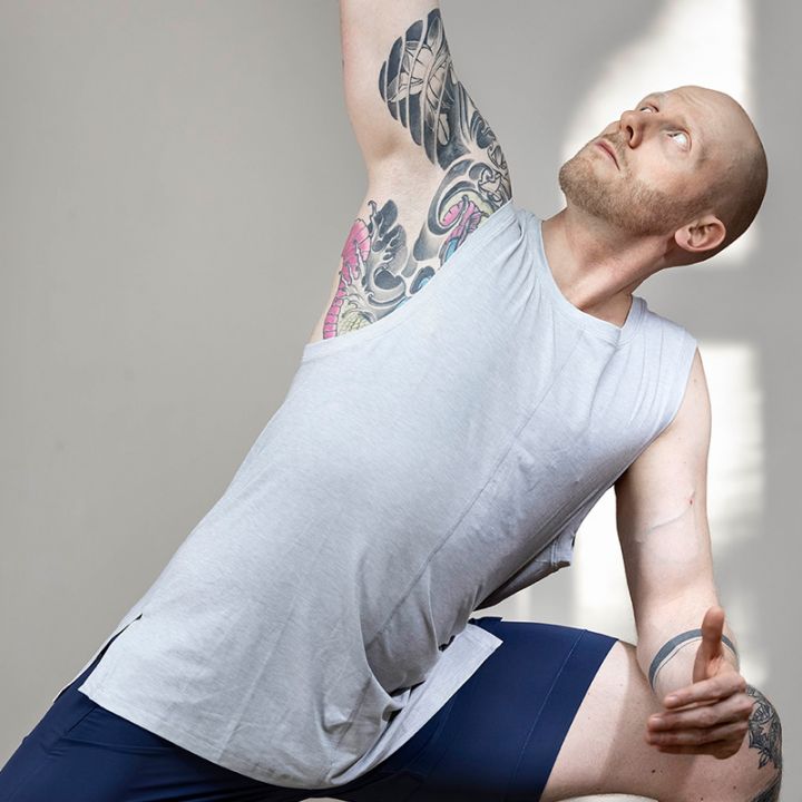 Eine Person mit Tattoos macht drinnen eine Yoga-Pose. Sie trägt ein graues ärmelloses Shirt und blaue Shorts. Sie blickt nach oben, ein Arm ist erhoben und der andere gebeugt, was eine dynamische und konzentrierte Haltung ergibt.