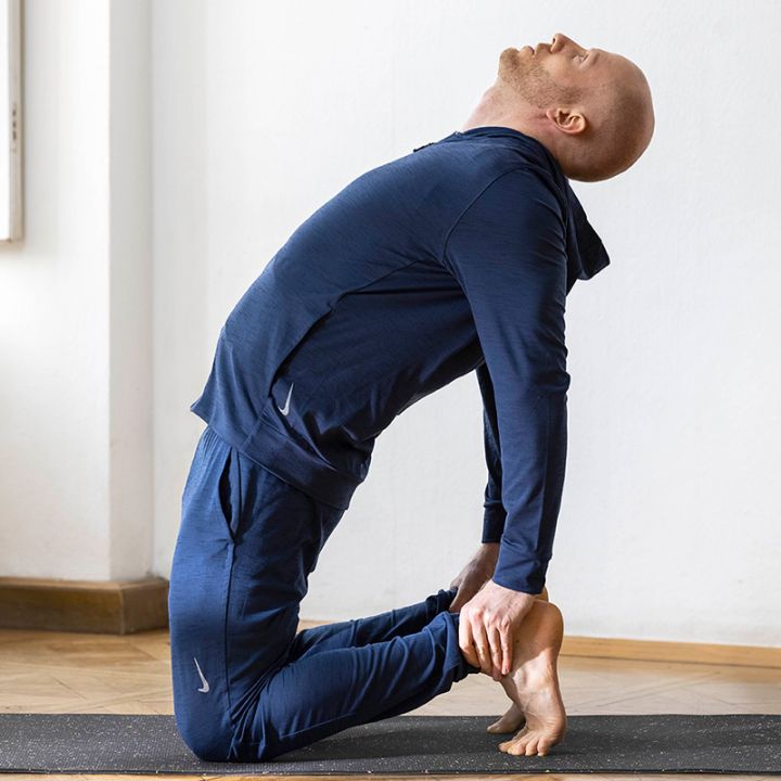 Ein Mann führt in einem hellen Raum auf einer Yogamatte eine Kamel-Yoga-Pose aus. Er trägt einen marineblauen Trainingsanzug, kniet mit gewölbtem Rücken und den Händen auf den Fersen und blickt nach oben. Im Hintergrund ist ein Fenster zu sehen.