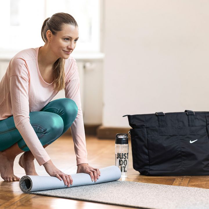 Eine Frau in einem hellrosa Oberteil und blaugrünen Leggings kniet und rollt eine Yogamatte zusammen. Neben ihr stehen eine schwarze Sporttasche und eine Wasserflasche. Sie steht drinnen auf einem Holzboden, im Hintergrund ist ein helles Fenster.