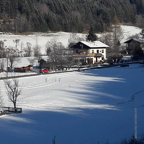 Winterlandschaft mit einem schneebedeckten Feld im Vordergrund. Ein paar Häuser und Bäume säumen die Mitte, auf der Straße ist ein rotes Auto zu sehen. Im Hintergrund sind schneebedeckte, bewaldete Hügel unter einem klaren Himmel zu sehen.