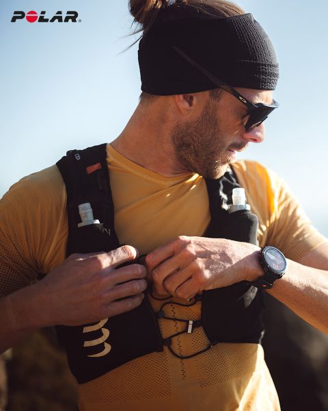 Eine Person mit schwarzem Stirnband und Sonnenbrille rückt ihre schwarze Trinkweste zurecht. Sie trägt ein gelbes Hemd und eine Smartwatch am Handgelenk. Das Polar-Logo ist oben links zu sehen. Sonnenlicht erhellt die Szene.