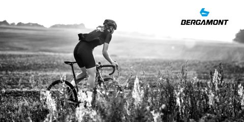 Ein Radfahrer mit Helm fährt mit einem Rennrad durch eine Wiese, im Hintergrund ist eine hügelige Landschaft zu sehen. Das Bild ist schwarzweiß und in der oberen rechten Ecke ist das Bergamont-Logo zu sehen.