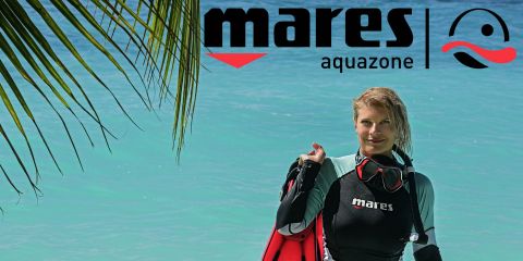 Eine Frau in einem schwarz-grünen Neoprenanzug mit dem „Mares“-Logo steht am türkisfarbenen Wasser und hält eine rote Taucherausrüstung in der Hand. Über ihr hängen Palmblätter. Das „Mares Aquazone“-Logo befindet sich in der oberen rechten Ecke.