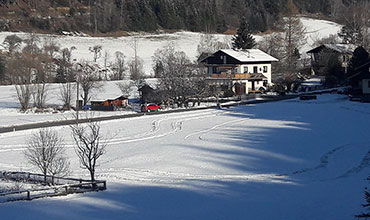 Eine verschneite Landschaft mit einem von Bäumen umgebenen Haus. Eine gewundene Spur aus Fußspuren durchquert das schneebedeckte Feld. Im Hintergrund sind vereinzelte Bäume und weitere Gebäude unter einem teilweise bewölkten Himmel zu sehen.