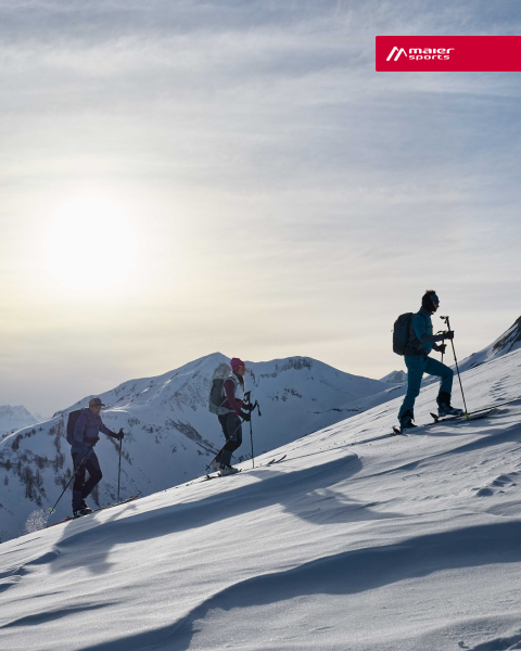 Drei Personen in Winterkleidung wandern unter strahlendem Sonnenschein einen schneebedeckten Berghang hinauf. Sie benutzen Skistöcke und tragen Rucksäcke. Im Hintergrund sind schneebedeckte Gipfel in der Ferne zu sehen.