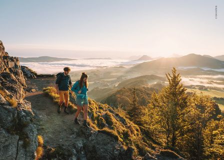 Zwei Wanderer, ein Mann und eine Frau, gehen einen felsigen Pfad entlang, von dem aus man ein Tal überblicken kann. Die Sonne geht hinter den Bergen in der Ferne unter und wirft einen warmen Schein auf eine Landschaft mit sanften Hügeln und vereinzelten Wolken. Unten sind Bäume und grüne Felder zu sehen.