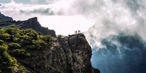 Eine Gruppe von Wanderern steht auf einer Felsklippe, deren Silhouette sich vor einem wolkigen Himmel abzeichnet. Die Landschaft zeichnet sich durch dramatische Felsformationen und viel Grün aus, im Hintergrund sind neblige Berge zu sehen.