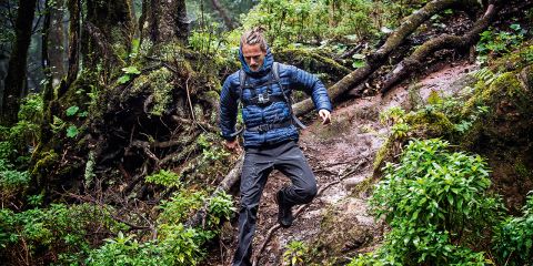 Eine Person in Outdoor-Kleidung navigiert einen schroffen, nassen Waldweg. Sie trägt eine Jacke und einen Rucksack und geht vorsichtig auf einem schlammigen Pfad, der von üppigem Grün und Baumwurzeln umgeben ist. Die Atmosphäre ist neblig und feucht.