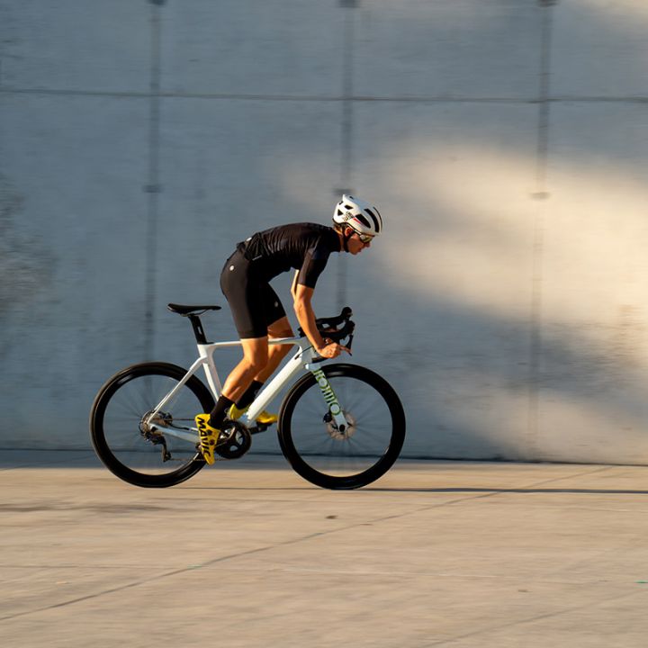Ein Radfahrer in schwarzem Outfit und weißem Helm fährt mit seinem weißen Fahrrad schnell an einer Betonwand vorbei. Boden und Wand haben eine glatte Textur und die Szene wird vom Sonnenlicht beleuchtet, das weiche Schatten wirft.