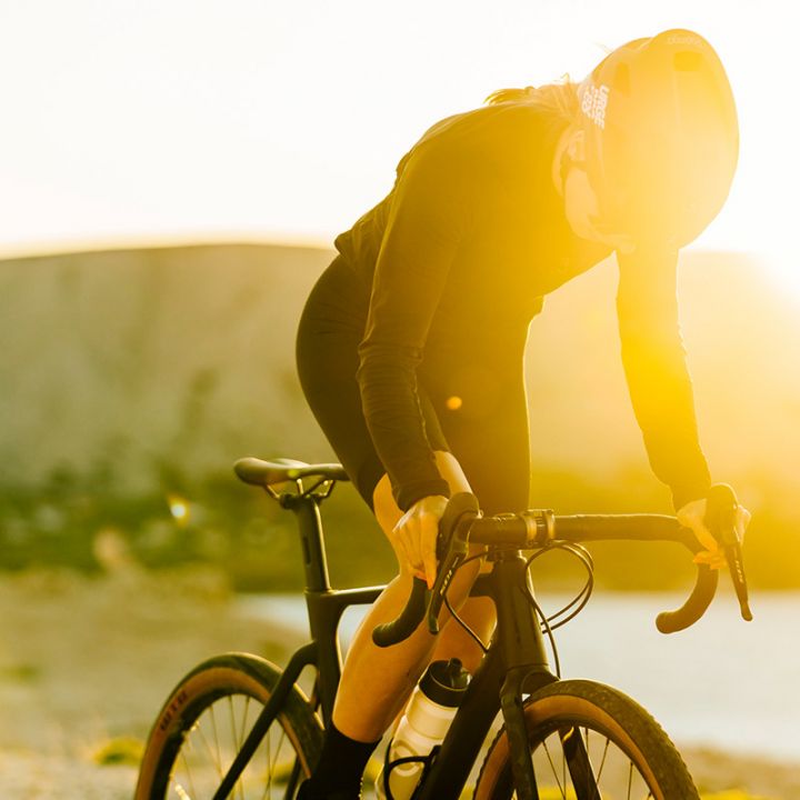 Ein Radfahrer mit Helm und dunkler Kleidung lehnt sich über sein Rennrad, während die helle Sonne einen warmen Schein erzeugt. Malerischer Hintergrund mit verschwommenen Klippen und Wasser, was an einen Standort im Freien bei Sonnenuntergang oder Sonnenaufgang erinnert.