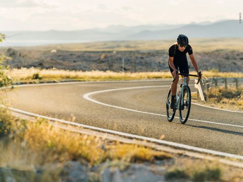 Radfahrer in schwarzer Ausrüstung fährt auf einer kurvenreichen Straße durch eine malerische, offene Landschaft mit Bergen in der Ferne unter einem teilweise bewölkten Himmel. Die Sonne wirft einen warmen Schein auf die Szene.