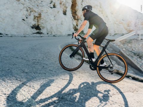 Ein Radfahrer in schwarzem Outfit und Helm fährt an einem sonnigen Tag eine kurvenreiche Bergstraße hinauf. Die Sonne wirft lange Schatten auf die Straße und im Hintergrund ist eine Felsklippe zu sehen.