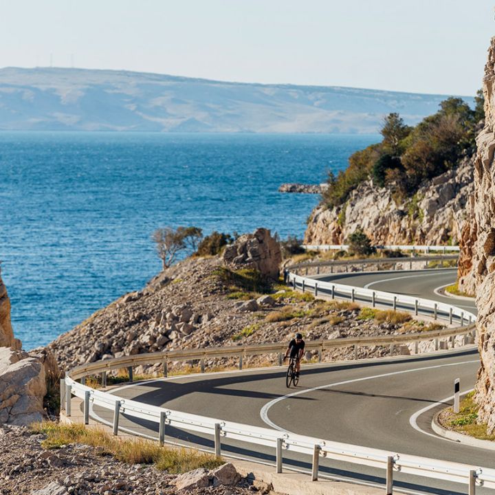 Ein Radfahrer fährt eine kurvenreiche Küstenstraße entlang, rechts von felsigen Klippen und links vom Meer. Am Horizont sind in der Ferne Hügel unter einem klaren blauen Himmel zu sehen.