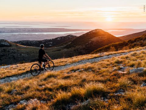 Ein Radfahrer in schwarzem Outfit fährt bei Sonnenuntergang mit seinem Mountainbike auf einem Grasweg. Die Szene bietet einen Blick auf sanfte Hügel und ein entferntes Gewässer unter einem farbenfrohen Himmel. Die Sonne wirft einen warmen Schein auf die Landschaft.