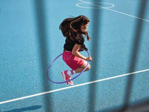 Ein junges Mädchen mit geflochtenen Haaren springt auf einem blauen Sportplatz durch einen lila Hula-Hoop-Reifen. Sie trägt ein schwarzes Shirt, rosa Shorts und rote Turnschuhe mit weißen Socken. Das Bild ist teilweise von unscharfen vertikalen Linien eingerahmt.